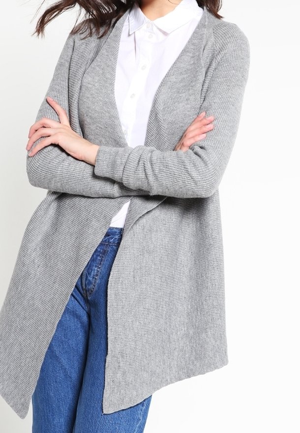 Harmaa avoin neulejakku, jossa on kuvioitu neulospinta, pitkät hihat ja laskeutuva malli. Yhdistetty valkoiseen paitaan ja sinisiin farkkuihin.