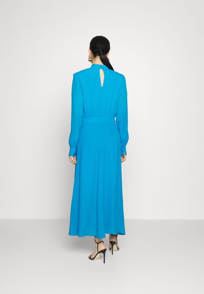 Długa rękaw, jasnoniebieska sukienka z wcięciem w pasie, szczegółem wycięcia na plecach oraz zwiewnym dołem. Zestawiona z czarnymi sandałami na wysokim obcasie.