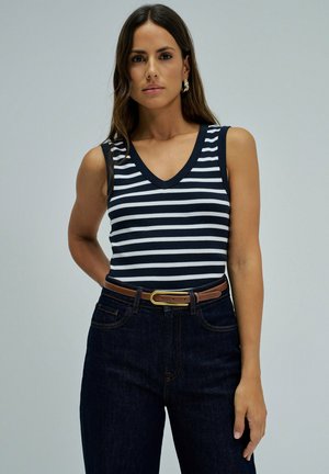Mujer con camiseta sin mangas a rayas negras y blancas, vaqueros oscuros con cinturón marrón, de pie con expresión neutral frente a un fondo liso.