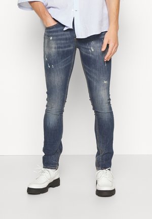 Hombre vistiendo jeans azul oscuro ajustados y desgastados, con una camisa azul clara y zapatos blancos con cordones, de pie frente a un fondo liso.