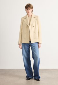 Béžový dvojradový vlnený kabát s vystrihnutým golierom, skombinovaný s svetlomodrými širokými nohavicami a čiernymi topánkami s ostrou špičkou.