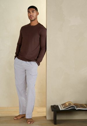 Muž sa opiera o béžovú stenu, oblečený v hnedej košeli s dlhým rukávom a bielych pruhovaných nohaviciach, bosý vedľa nízkeho stola s otvorenou knihou.