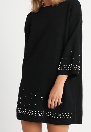 Femme portant une robe noire ample à manches trois-quarts, décorée de clous argentés sur les manches et l'ourlet.