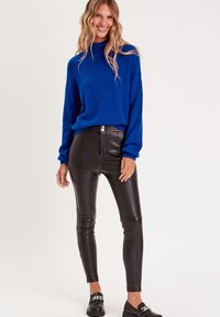 Blauer strukturierter Pullover mit langen Ärmeln, kombiniert mit glänzenden schwarzen Kunstlederhosen und schwarzen Slippern mit Kettendetail.