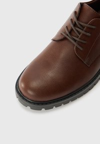 Zapato de vestir de cuero marrón con superficie texturizada, punta redondeada, cordones grises y suela de goma negra con patrón de banda.