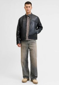 Giubbotto di pelle nera con colletto alto, tasca con zip, camicia a quadri sotto, abbinato a jeans grigi sbiaditi e calzature beige.