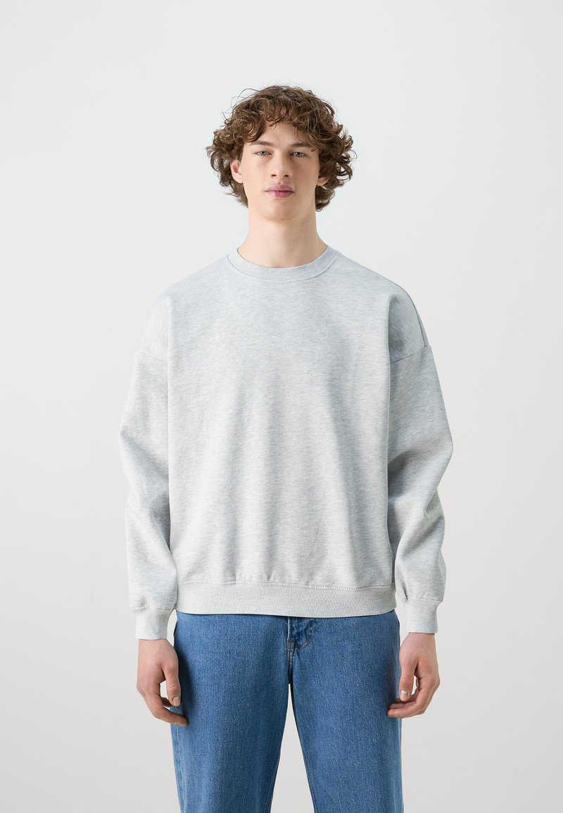 Felpa grigia chiaro con collo rotondo, polsini e fondo a costine. Abbinata a jeans blu con un design a vestibilità rilassata.