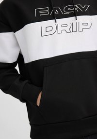 Sudadera en blanco y negro con un diseño texturizado, un bolsillo canguro central y el texto impreso "EASY DRIP" en la parte frontal.
