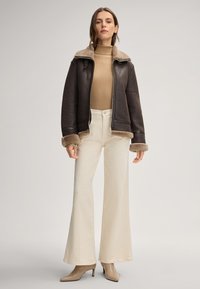 Braune Lederjacke mit Schaffellkragen und -manschetten, kombiniert mit einem beigen gerippten Rollkragenpullover und cremefarbener, weit geschnittener Hose. Beigefarbene Schuhe mit Absatz.