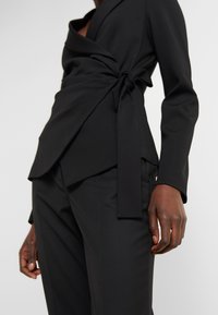 Svart omslagstopp med lång ärm, med en knytning i midjan och en mjuk tygtextur. Parat med skräddarsydda svarta byxor.