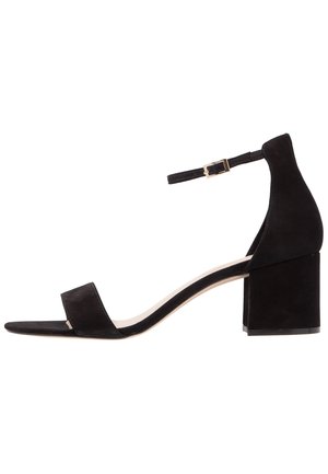 Sort ruskind højhælet sandal med blokhæl, åben tå, ankelrem og spænde i guldton. Har en glat tekstur og minimalistisk design.