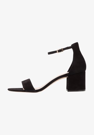 Black suede heeled sandal with a block heel, open toe, ankle strap, and gold-tone buckle. Features a smooth texture and minimalist design.