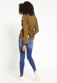 Olivgrüne Jacke mit verstellbarem Bund, langen Ärmeln und Bündchen. Kombiniert mit eng anliegenden blauen Jeans und weißen Sneakers, von hinten betrachtet.
