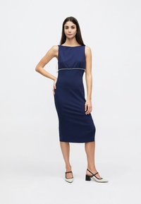 Femme debout dans une robe midi sans manches bleu marine avec bordure en perles, portant des chaussures à talons blocs blancs avec des lanières noires sur fond blanc.