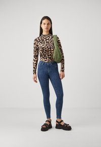 Žena stoji odjevena u top s uzorkom leopard, plave uske traperice, crne platforme s sandalama i nosi zeleni kviltirani rameni torbu.