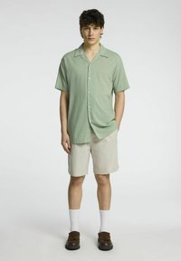 Camicia verde chiaro a maniche corte con colletto, abbinata a pantaloni corti beige. Il modello indossa calze bianche e scarpe marroni. Design semplice e rilassato.