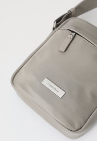Szara torebka typu crossbody Calvin Klein, wykonana z gładkiego materiału, z zamknięciem na zamek błyskawiczny, płaskim paskiem oraz metalową naszywką z logo na przodzie.