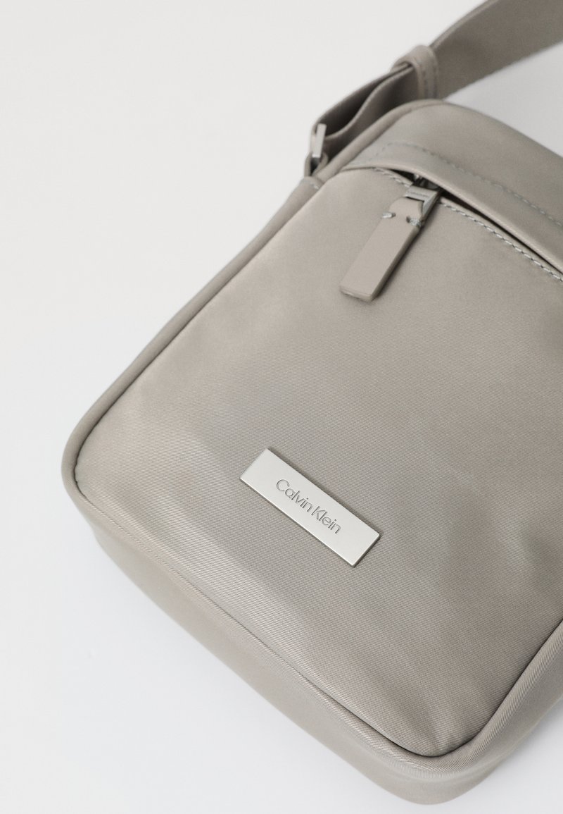 Borsa a tracolla grigia Calvin Klein realizzata in tessuto morbido, dotata di chiusura con zip, tracolla piatta e una placca logo in metallo sulla parte anteriore.