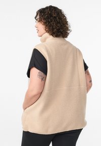 Beige fleece vest met hoge kraag, mouwloos ontwerp en gestructureerd oppervlak. Heeft zijzakken en wordt gedragen over een zwarte top.
