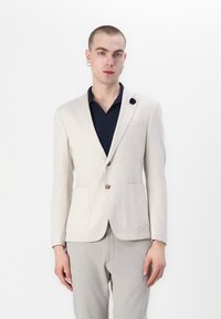 Lys beige skreddersydd blazer med to frontlommer, brune knapper og en marineblå skjorte under. Glatt stoff og rene linjer.