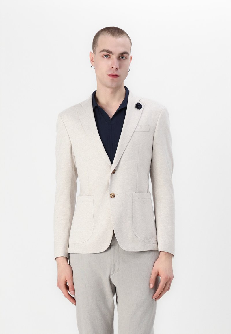 Lys beige skreddersydd blazer med to frontlommer, brune knapper og en marineblå skjorte under. Glatt stoff og rene linjer.