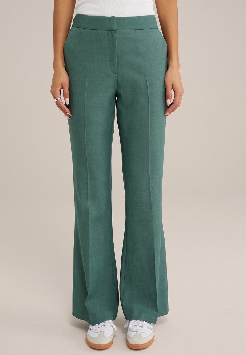 Gröna utsvängda byxor med en slät textur, hög midja, framfåra och sidofickor, parat med vita sneakers med bruna sulor.