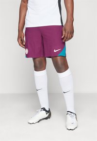 Pantaloni da calcio Nike di colore bordeaux con accenti teal, caratterizzati da un tessuto testurizzato, un logo cucito e una striscia laterale. Abbinati a calze bianche e scarpe da calcio.