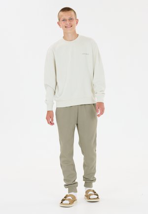 Ung mand står og smiler, iført en hvid sweatshirt, beige joggingbukser, hvide sokker og brune sandaler mod en hvid baggrund.