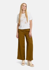 T-shirt blanc à manches courtes avec broderie florale en or et beige, associé à un pantalon taille haute à jambes larges de couleur olive et des talons épais de couleur claire.
