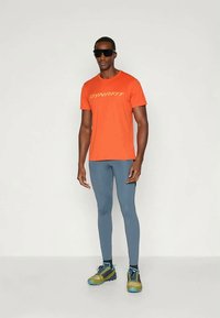 Oranž T-särk lühikeste varrukatega, millel on "DYNAFIT" logo, kombineeritud siniste retuusidega ja mitmevärviliste matkasussidega. Mudel kannab prille.