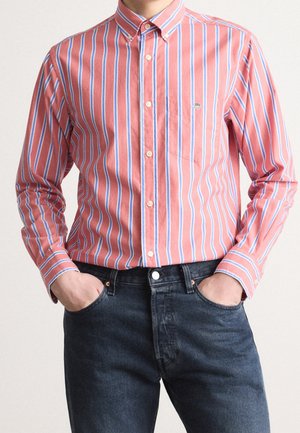 Mężczyzna ubrany w różową koszulę z długim rękawem w pionowe białe i niebieskie paski, włożoną do ciemnoniebieskich jeansów, z rękami w kieszeniach, stojący na jednolitym tle.