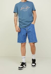 Camiseta azul con logo blanco y rosa, combinada con pantalones cortos de mezclilla azules. Zapatillas negras y blancas, calcetines blancos con nombre de la marca. Fondo claro.