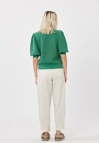 Femme aux cheveux blonds ondulés portant un haut en maille vert à manches bouffantes, un pantalon blanc ample et des sandales beiges, debout dos à un fond blanc.
