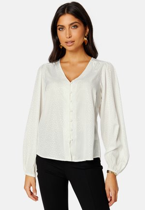 Blusa bianca con scollatura a V, maniche lunghe a palloncino e chiusure a bottoni. Presenta un sottile motivo testurizzato e un tessuto liscio simile al satin.