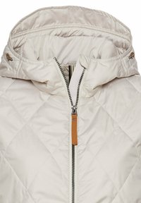 Veste matelassée beige clair avec col montant, fermeture éclair à l'avant avec tirette marron et capuche réglable avec cordons de serrage.