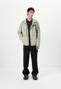 Light green hooded jacket with large front pockets, lightweight fabric, and a relaxed fit, paired with black pants and chunky black sneakers.