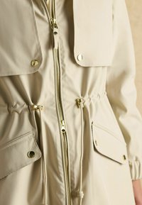 Bežs ūdensizturīgs mētelis ar zipa priekšējo daļu, elastīgu vidukli, lielām kabatām ar atloku un zelta toņu apdari. Gluda tekstūra un aukliņas detalizācija.