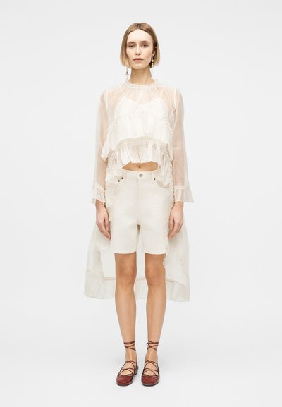 Femme portant un haut blanc transparent à manches longues avec volants et ourlet dégradé, short blanc, et des ballerines bordeaux à lacets, debout devant un fond blanc.