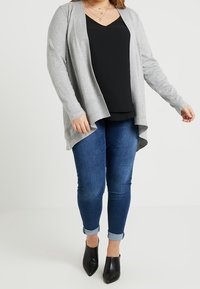 Cárdigan gris claro con frente abierto, mangas largas y puños acanalados, superpuesto a una camiseta negra y combinado con unos vaqueros azules y tacones negros.
