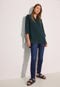 Tunica lunga a maniche lunghe di colore verde scuro con collo a bottoni; abbinata a jeans slim-fit blu scuro e sandali navy con suola in sughero.