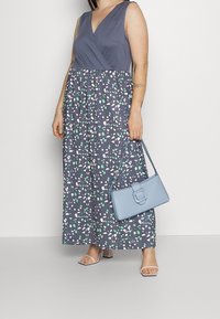 Maxi šaty v tmavě šedé barvě s bílým a zeleným abstraktním potiskem, výstřih do V, bez rukávů, doplněné světle modrou kabelkou a bílými podpatky.