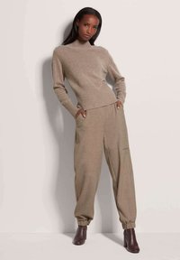 Beige Strickpullover mit Stehkragen und geripptem Saum, kombiniert mit lockeren beigen Hosen mit Seitentaschen und elastischen Bündchen; braune Stiefeletten mit Absatz.
