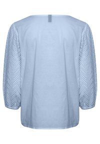 Ljusblå blus med en slät kropp och texturerade, mönstrade öglaärmar som har chevronmönster. Detalj på bakre halsknäppning synlig.