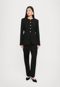 Zwarte suit met een op maat gemaakte blazer met gouden knopen en twee voorzakken, gecombineerd met rechte broekspijpen en puntenballetjes.