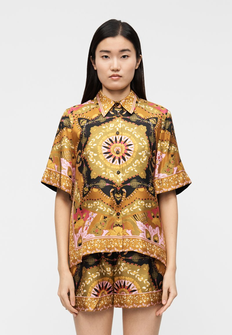 Short-sleeve, button-up shirt in golden fabric featuring bold floral and geometric patterns in pink, black, and green. Smooth texture.