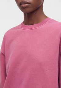 Gros plan d'une personne portant un sweat-shirt rose uni à col rond, montrant le cou, le bas du visage et le haut du torse sur un fond blanc.