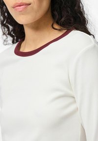 Kvinna med lockigt mörkt hår, iförd en vit ribbad tröja med en mörkröd krage, visad från halsen till övre bröstet.