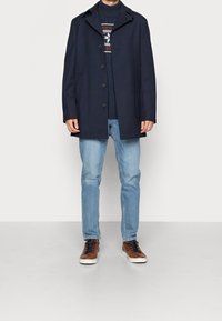 Marineblå ullblandad kappa med snedskuren krage, fem knappar, stylad över en mörk tröja, kombinerad med ljusblå jeans och bruna sneakers.