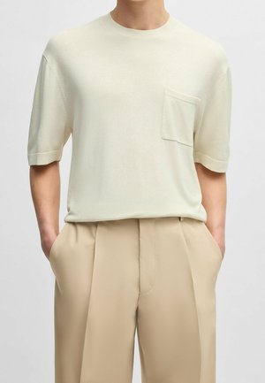 Jasno beżowa koszulka z krótkim rękawem, okrągłym dekoltem i małą kieszonką na piersi. Miękka, gładka faktura. Połączona z beżowymi spodniami w kant.