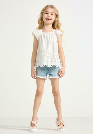 Ung pige med langt blondt hår iført en hvid broderet top, denimshorts med blondekant og blomstrede sløjfesko, står og smiler.